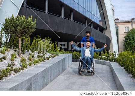 Handsome man sitting in a wheelchair while having a walk with a male nurse Handsome man sitting in a wheelchair while having a walk with a male nurse 112698804