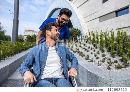 Handsome man sitting in a wheelchair while having a walk with a male nurse 112698813