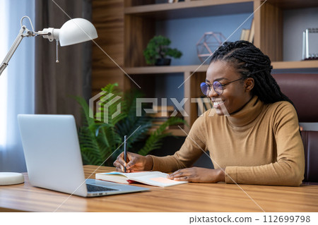 Woman in glasses with dreadlocks sitting at workplace with laptop writing in her paper notebook 112699798