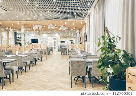 Side view of a large empty restaurant with tables, soft chairs, spotlights on ceiling, wooden floor and plant. Modern interior of a cafe, restaurant for events, holidays, weddings with copy space. 112700301