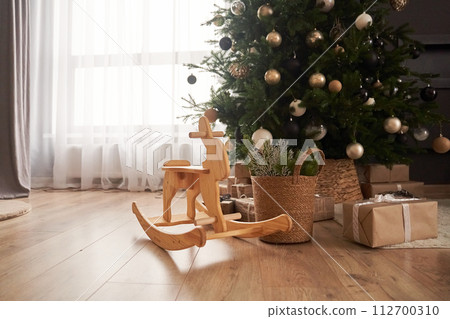 View of wooden rocking rabbit in the center of living room with decorated christmas tree, wicker basket with spruce branches, gifts in craft paper with copy space. View of wooden rocking rabbit in the center of living room with decorated christmas tree, wicker basket with spruce branches, gifts in craft paper with copy space. 112700310