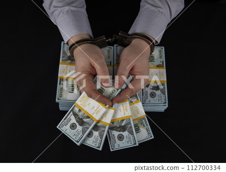 An arrested male official in handcuffs holds stacks of dollars in his hands received as a bribe. A dishonest official is arrested while accepting a bribe. Handcuffs on hands and dollars. An arrested male official in handcuffs holds stacks of dollars in his hands received as a bribe. A dishonest official is arrested while accepting a bribe. Handcuffs on hands and dollars. 112700334