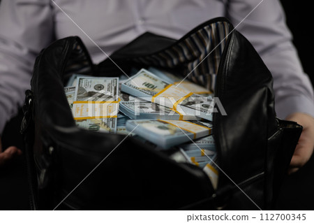 Close-up of an unzipped bag full of stacks of hundred dollar bills in front of a businessman on a black background. New packs of dollars lie in a black bag close-up. Concept of big money, victory. 112700345