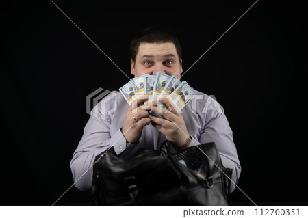 An official with joyful eyes inhales the smell of money in his hands on a black background. A man in joyful amazement holds a fan of stacks of dollars in front of his face. An official with joyful eyes inhales the smell of money in his hands on a black background. A man in joyful amazement holds a fan of stacks of dollars in front of his face. 112700351