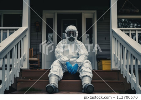 A disinfection service worker in a protective suit and gas mask sits on the porch of the house 112700570
