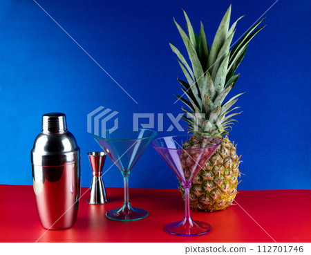 Cocktail Shaker with Cocktail Glasses and Pineapple on a Red Base and Blue Background 112701746
