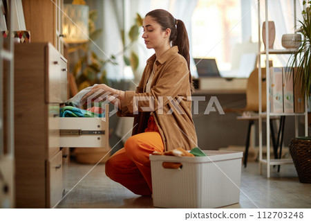 Side view portrait of young woman storing clean clothes in drawers and enjoying laundry day, copy space Side view portrait of young woman storing clean clothes in drawers and enjoying laundry day, copy space 112703248