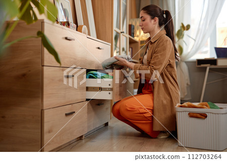 Full length side view portrait of young woman putting away clean clothes in drawer on laundry day, copy space 112703264