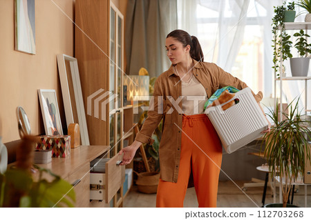 Side view portrait of young woman putting away clean clothes on laundry day, copy space 112703268
