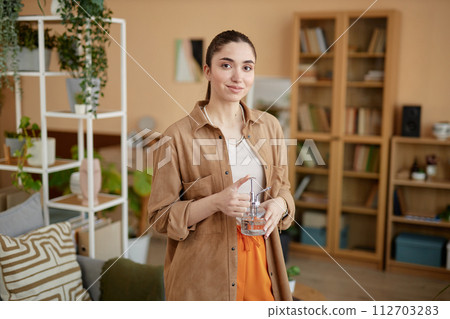 Waist up portrait of young woman holding glass watering can and smiling at camera at home copy space 112703283