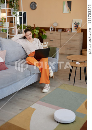 Vertical full length portrait of young woman using laptop on couch and relaxing with robot vacuum cleaner mopping floors Vertical full length portrait of young woman using laptop on couch and relaxing with robot vacuum cleaner mopping floors 112703293