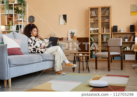 Wide angle view at young woman relaxing on couch with smart robot vacuum cleaner mopping floors copy space 112703296