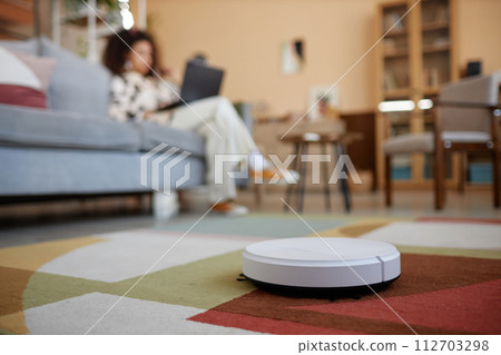 Close up of white robot vacuum cleaner on colorful carpet with blurred young woman in background copy space Close up of white robot vacuum cleaner on colorful carpet with blurred young woman in background copy space 112703298