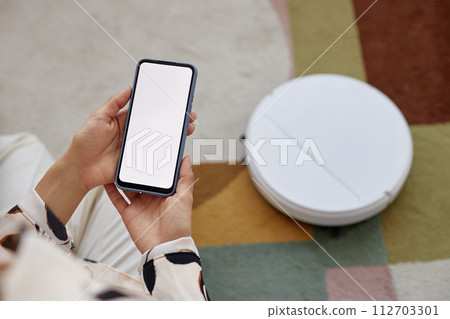 Top view closeup of unrecognizable woman holding smartphone with blank white screen while operating robot vacuum cleaner at home copy space 112703301