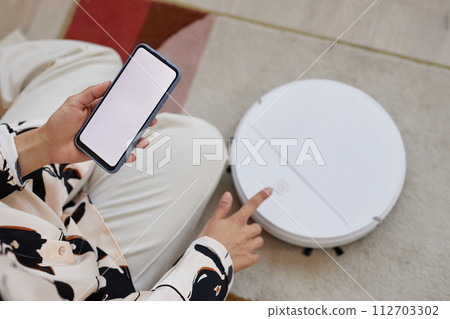 Top view closeup of unrecognizable young woman operating smart robot vacuum cleaner and holding smartphone with blank white screen copy space 112703302
