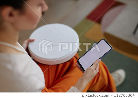 High angle closeup of young woman setting up robot vacuum cleaner and holding smartphone with white blank screen 112703304