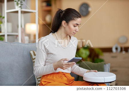 Side view portrait of young woman setting up robot vacuum cleaner and connecting to smartphone copy space 112703306