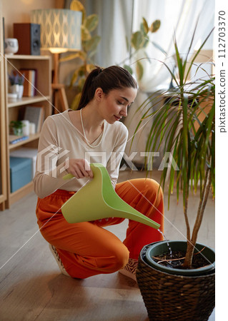 Vertical full length portrait of smiling young woman watering potted palm tree houseplant in cozy home Vertical full length portrait of smiling young woman watering potted palm tree houseplant in cozy home 112703370