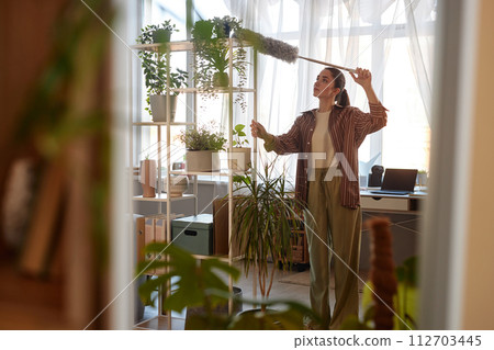 Mirror reflection of young woman dusting tall shelves with live green plants while refreshing house copy space 112703445