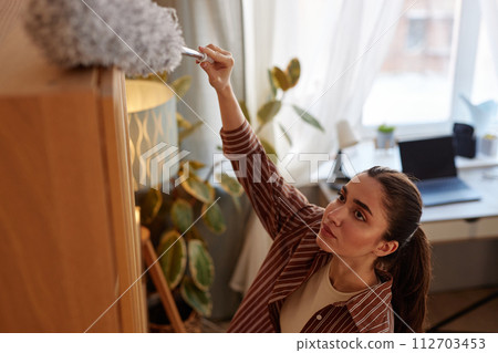 High angle portrait of young woman dusting tall shelves and hard to reach places while doing Spring cleaning at home 112703453
