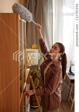 Vertical portrait of young woman dusting top of tall cupboard while doing Spring cleaning at home Vertical portrait of young woman dusting top of tall cupboard while doing Spring cleaning at home 112703456