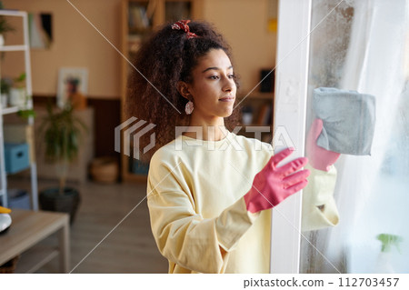 Side view portrait of young African American woman with curly hair cleaning windows at home and wearing pink rubber gloves copy space Side view portrait of young African American woman with curly hair cleaning windows at home and wearing pink rubber gloves copy space 112703457