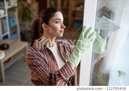 Side view portrait of young woman cleaning windows at home and wearing rubber gloves copy space Side view portrait of young woman cleaning windows at home and wearing rubber gloves copy space 112703458