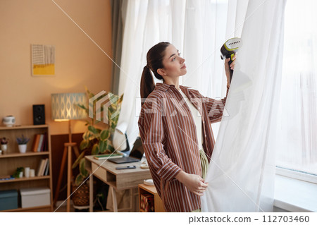 Side view portrait of adult woman enjoying Spring cleaning at home and steaming curtains on window copy space Side view portrait of adult woman enjoying Spring cleaning at home and steaming curtains on window copy space 112703460