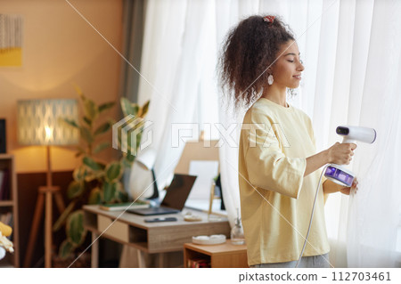 Wide angle side view at smiling young woman enjoying Spring cleaning and steaming curtains on window at home copy space Wide angle side view at smiling young woman enjoying Spring cleaning and steaming curtains on window at home copy space 112703461