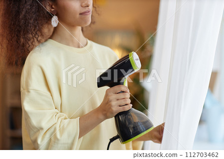 Close up of unrecognizable young woman holding hand steamer appliance and steaming curtains on window copy space Close up of unrecognizable young woman holding hand steamer appliance and steaming curtains on window copy space 112703462