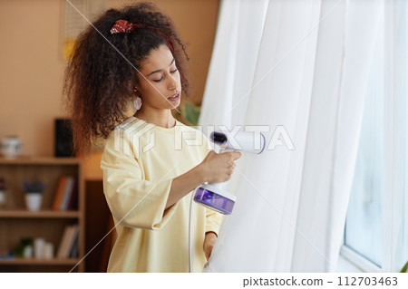 Waist up portrait of young woman steaming gauze curtains on window with with hand steamer during Spring cleaning copy space 112703463