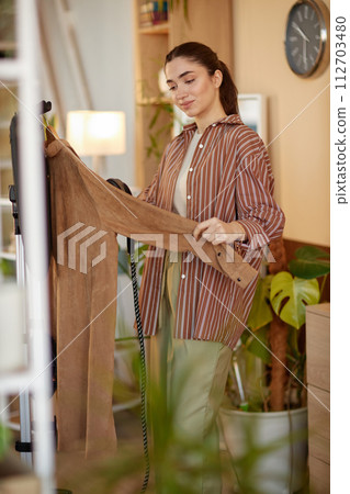 Vertical portrait of smiling young woman taking care of delicate clothes and using garment steamer at home 112703480