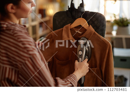 Close up of young woman using garment steamer on vertical ironing board and steaming suede jacket copy space Close up of young woman using garment steamer on vertical ironing board and steaming suede jacket copy space 112703481