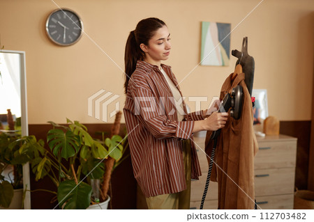 Side view portrait of young woman using garment steamer on velvet jacket in cozy home copy space 112703482