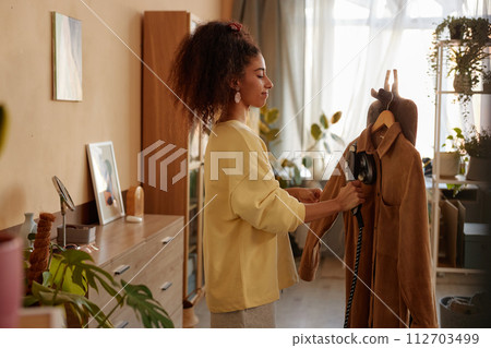 Side view portrait of young woman with curly hair steaming velvet jacket while taking care of clothes in cozy home Side view portrait of young woman with curly hair steaming velvet jacket while taking care of clothes in cozy home 112703499