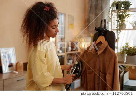 Side view portrait of curly haired young woman steaming delicate velvet clothing at home copy space Side view portrait of curly haired young woman steaming delicate velvet clothing at home copy space 112703501
