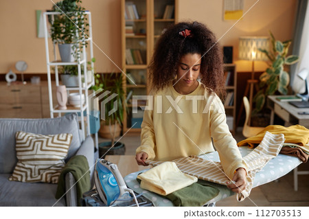 High angle portrait of curly haired young woman ironing and folding clothes on laundry day and taking care of house chores copy space 112703513