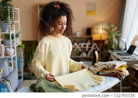 Waist up portrait of smiling young woman with big curly hair folding clothes on ironing board at home copy space 112703516