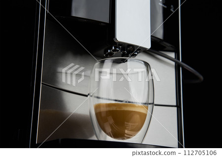 Coffee machine on dark background, close up. Modern coffee maker with freshly brewed coffee in glass cup. Kitchen appliances Coffee machine on dark background, close up. Modern coffee maker with freshly brewed coffee in glass cup. Kitchen appliances 112705106
