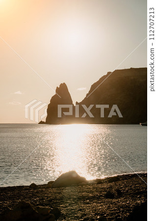 A red burning sunset over the sea with rocky volcanic cliff. Abstract nature summer ocean sea background. Small waves on golden warm water surface with bokeh lights from sun. 112707213
