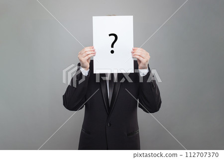 Business man wearing black suit hiding his face behind white paper with black question mark 112707370