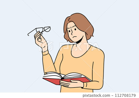 Woman experiences vision problems reading book and looks at glasses with upset grimace 112708179