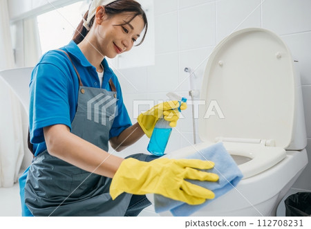 A woman wearing gloves scrubs toilet seat with cloth emphasizing purity in bathroom cleaning. Her meticulous work reflects dedication to housekeeping and hygiene. Housekeeper healthcare concept 112708231