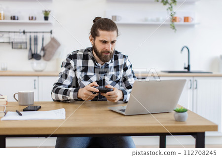 Caucasian man using joystick and laptop sitting at wooden table at kitchen. 112708245