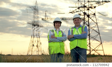 Happy workers team stands crossing arms against electric power transmission lines supports in sunset field. Engineers with crossed arms rest after work at power distribution substation at sunset Happy workers team stands crossing arms against electric power transmission lines supports in sunset field. Engineers with crossed arms rest after work at power distribution substation at sunset 112709942
