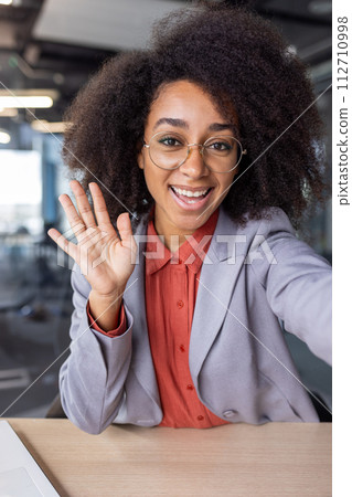 First person view on hispanic lady in trendy formalwear waving head for greeting gesture during video call. Friendly female in round eyeglasses joining conference with friend during working day. First person view on hispanic lady in trendy formalwear waving head for greeting gesture during video call. Friendly female in round eyeglasses joining conference with friend during working day. 112710998