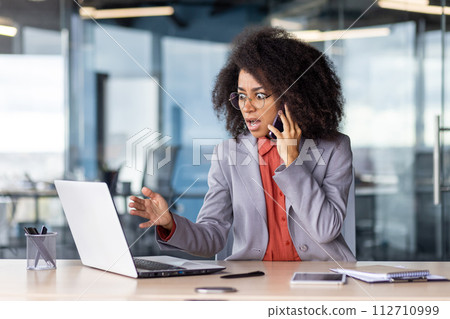 Terrified female talking by mobile phone while looking at portable computer screen with rounded eyes. Furious black woman getting desperate while receiving bad urgent news by business partner. 112710999