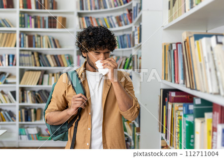 Unhealthy man in casualwear using paper napkin while sneezing and standing by aisle with collection of novels. Sick islamic male struggling with dust allergy in book shop while holding shoulder bag. Unhealthy man in casualwear using paper napkin while sneezing and standing by aisle with collection of novels. Sick islamic male struggling with dust allergy in book shop while holding shoulder bag. 112711004