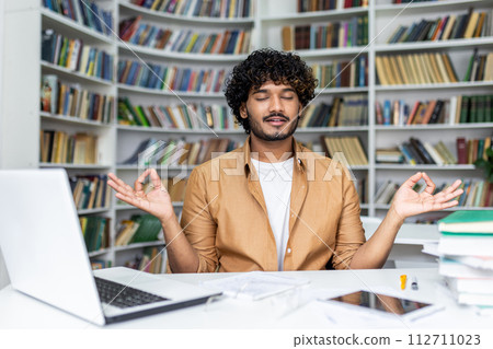 Middle eastern man sitting by desktop with computer and keeping raised hands in gyan mudra with closed eyes. Calm programmer using yoga pose practising for getting rid of stress and freeing mind. Middle eastern man sitting by desktop with computer and keeping raised hands in gyan mudra with closed eyes. Calm programmer using yoga pose practising for getting rid of stress and freeing mind. 112711023