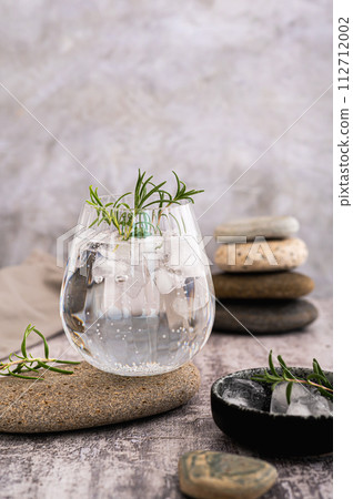 Gin tonic with ice and rosemary in a glass on a stone on the table vertical view Gin tonic with ice and rosemary in a glass on a stone on the table vertical view 112712002